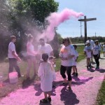 A group of people stand in a park on the sidewalk. There is pink dust in the air and on the sidewalk.