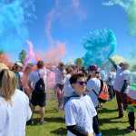 A group of people stand in a park on a sunny day, most wearing white shirts. Bursts of coloured dust fill the air in blues, greens and pinks.