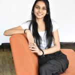 A woman with long, straight dark hair sits sideways on an orange couch and rests against the back of it. One of her arms is on the back of the couch and the other meets it with her fingers intertwined. She is wearing a white shirt and dark grey pants and several bracelets on each wrist.