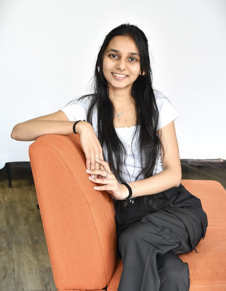 A woman with long, straight dark hair sits sideways on an orange couch and rests against the back of it. One of her arms is on the back of the couch and the other meets it with her fingers intertwined. She is wearing a white shirt and dark grey pants and several bracelets on each wrist.