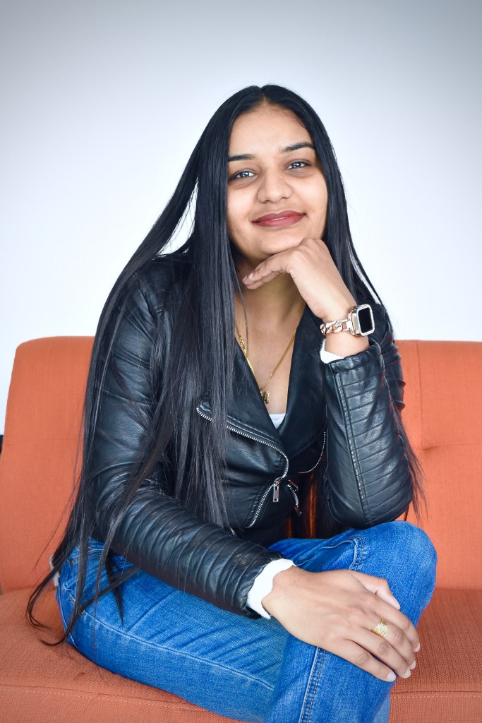a woman with long dark hair sits facing the camera on an orange couch. She is wearing a black leather jacket and blue jeans. Her legs are crossed and one arm is resting on her legs, supporting an arm so that her hand is up to support her chin. She is smiling at the camera.