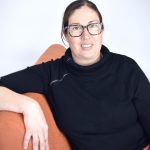 A woman with tied back brown hair and tortoise-shell glasses sits on an orange couch smiling naturally. She is sideways on the couch so that one arm rests on the back while the other sits beside her. She is wearing a black turtle neck and red pants.