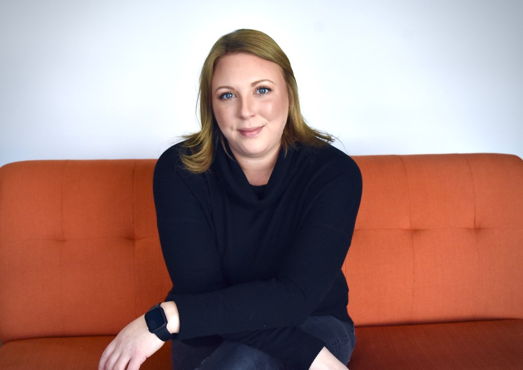A woman with dark blond hair and blue eyes sits on an orange couch facing the camera. She is wearing dark clothing and a smart watch and is smiling slightly. Her arms are crossed at the forearms and are resting on her legs.