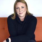 A woman with dark blond hair and blue eyes sits on an orange couch facing the camera. She is wearing dark clothing and a smart watch and is smiling slightly. Her arms are crossed at the forearms and are resting on her legs.