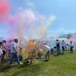 A large group of people stand in a park. Pink, green, red, blue and orange dye fill the air.
