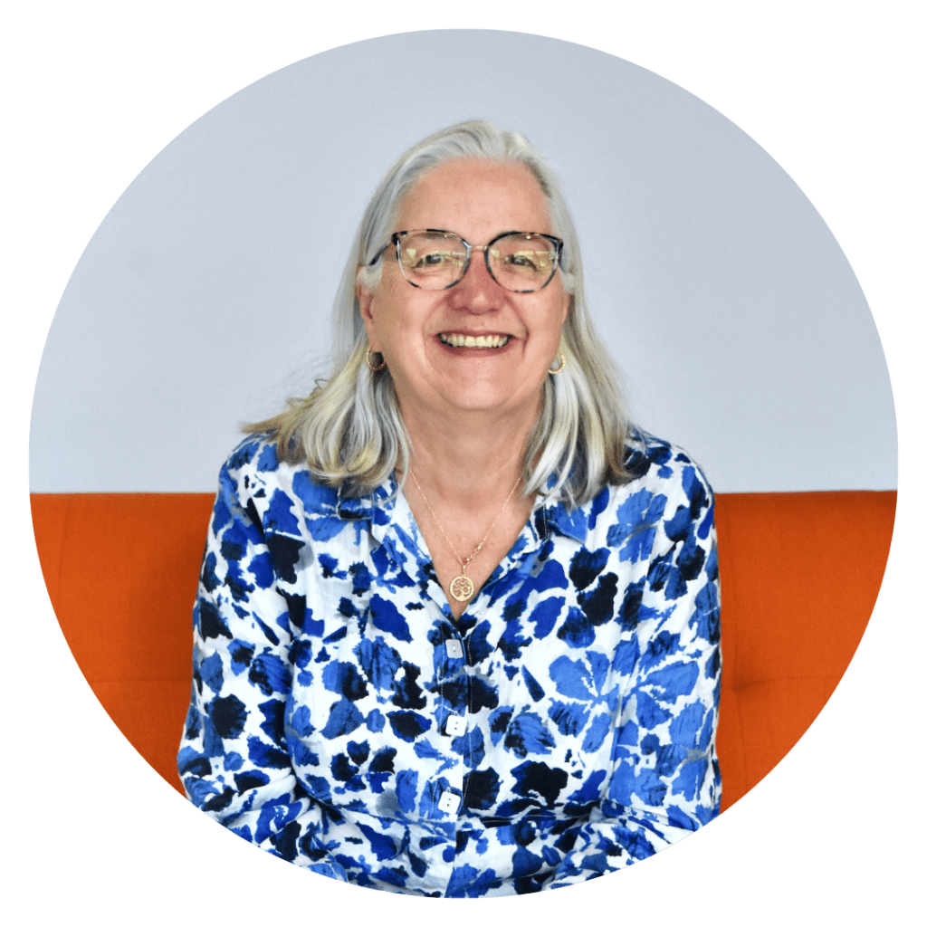 A woman with white hair and glasses sits on an orange couch smiling at the camera. She is wearing a blue and white floral shirt.