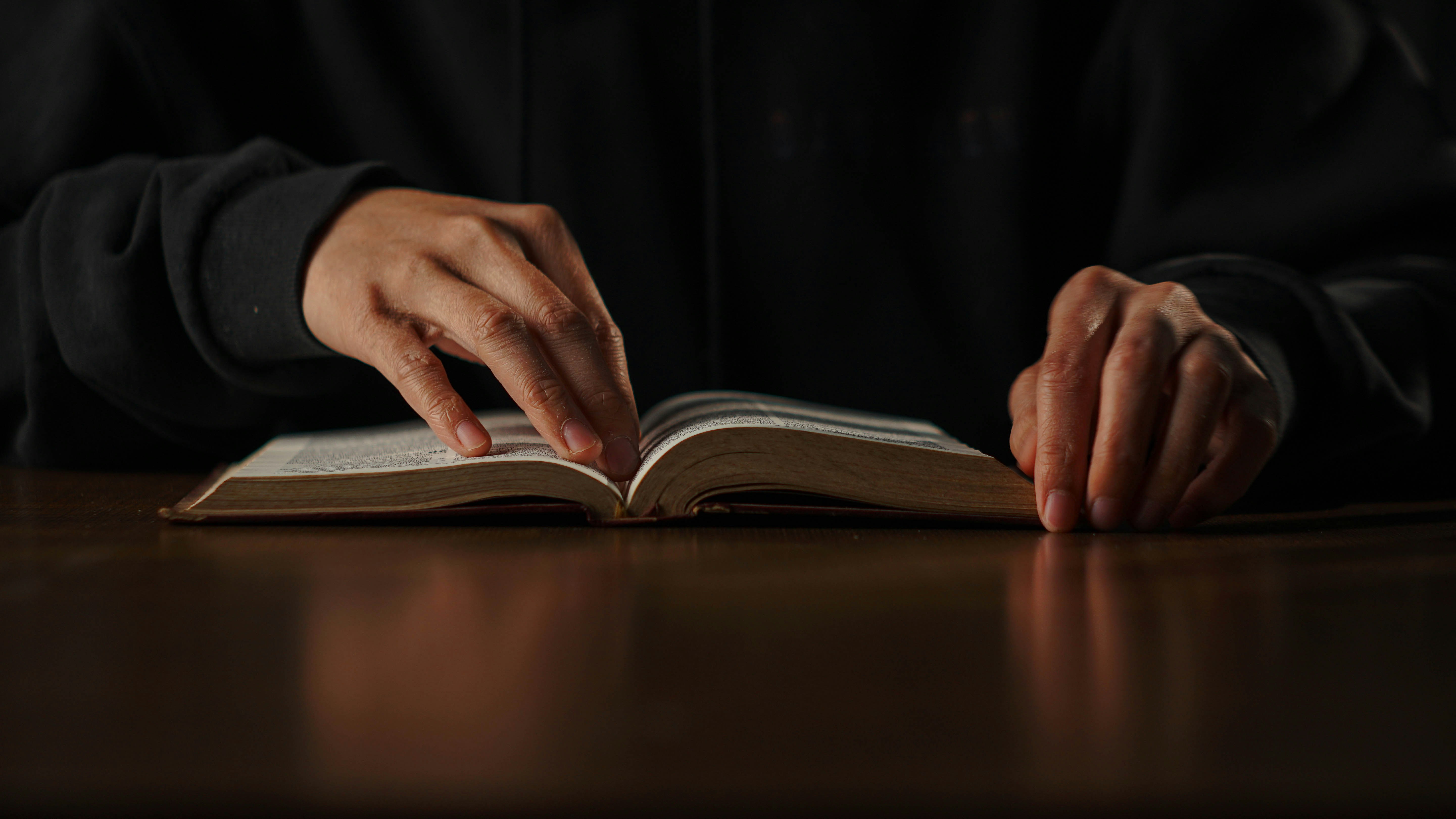 A book sits open on a table and two hands are shown. One hand props the book open while the other rests beside it.